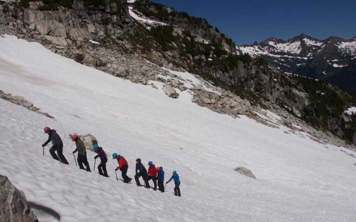 A group of people wearing mountaineering gear hike in a line up a snowy incline